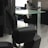 A barbershop or salon setting with black leather chairs and mirrors. The interior features a modern design with sleek lines and minimalist décor, including glass shelves and dark wooden cabinets. The room appears tidy and well-organized, creating a professional and upscale atmosphere.